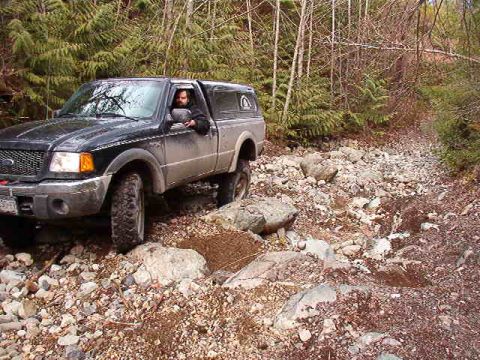 1985 Ford Bronco - A black Ford Ranger pickup truck with a camper shell is navigating a rocky, wooded trail. The driver is visible behind the wheel, looking f…