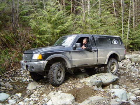 1985 Ford Bronco - A black Ford Ranger pickup truck with a black camper shell is navigating a rocky, wooded terrain. The driver is visible through the open dr…