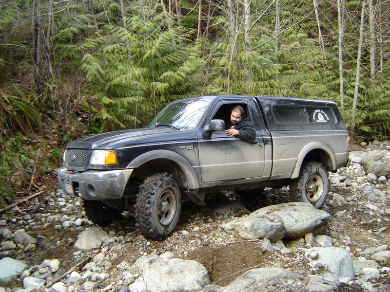 Black Ford Ranger pickup truck with camper shell driving on a rocky, wooded trail. Driver visible.