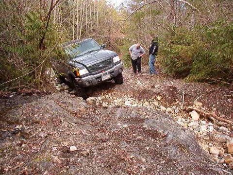 1985 Ford Bronco - A dark-colored Ford pickup truck is navigating a rocky, wooded trail. Two people stand nearby, observing the truck as it traverses the unev…