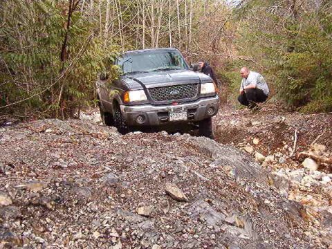 1985 Ford Bronco - A dark colored Ford Ranger pickup truck is navigating a rough, rocky terrain with two people nearby. The truck is angled on a steep incline…