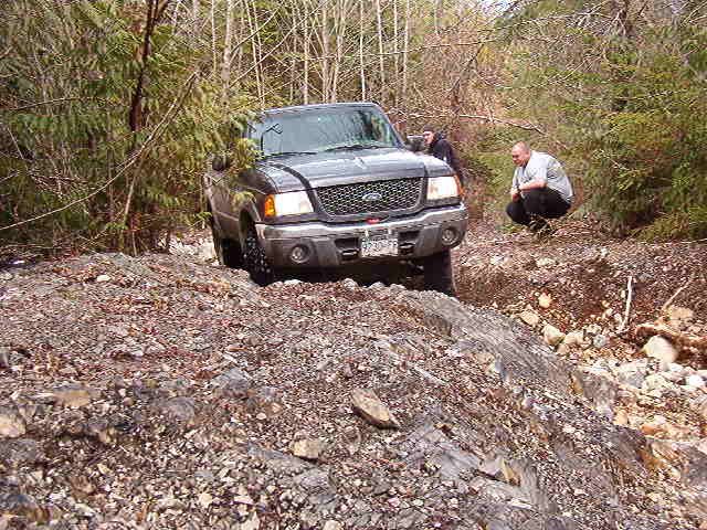 Dark Ford Ranger pickup truck on a rocky, off-road trail with two people watching.