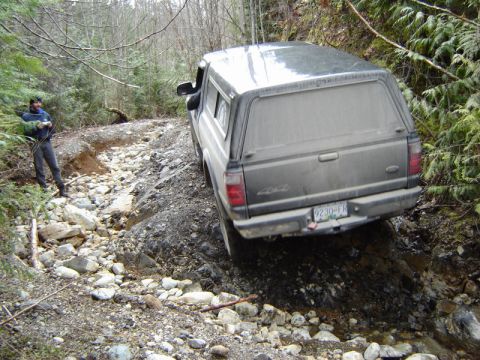 1985 Ford Bronco - A dusty, dark-colored pickup truck with a camper shell is navigating a rocky, off-road trail in a wooded area. A person stands on the left…
