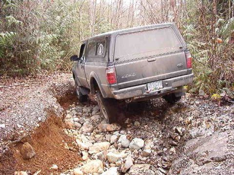 1985 Ford Bronco - A dark-colored pickup truck with a camper shell navigates a rough, rocky terrain with a steep ditch on the left side. The truck is angled,…