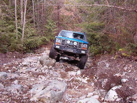 1985 Ford Bronco - A blue 1985 Ford Bronco is navigating a rocky, wooded trail with its front tires angled to the right. The off-road vehicle is positioned on…