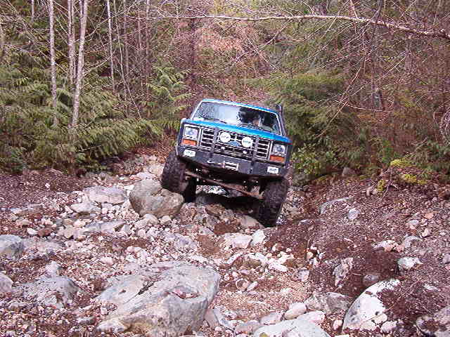 Blue 1985 Ford Bronco off-roading on a rocky, wooded trail.