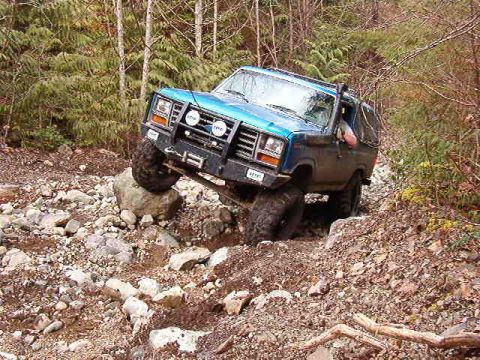 1985 Ford Bronco - A blue 1980s Ford Bronco is navigating a rocky, wooded trail with its front tire on top of a large rock. The off-road vehicle is tilted to…