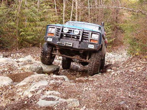 1985 Ford Bronco - A blue 1985 Ford Bronco is navigating a rocky, wooded trail, with its front tire on top of a large rock. The vehicle has a modified front b…