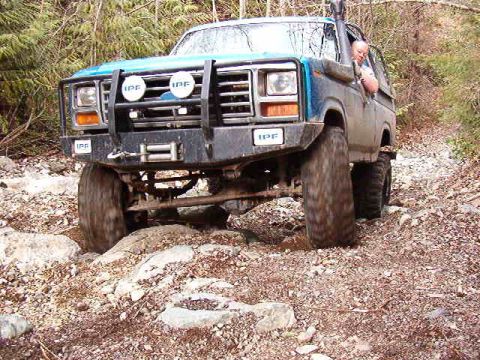 1985 Ford Bronco - A blue 1980s Ford Bronco is navigating a rocky, wooded trail. The off-road vehicle features a brush guard with IPF lights and a snorkel.