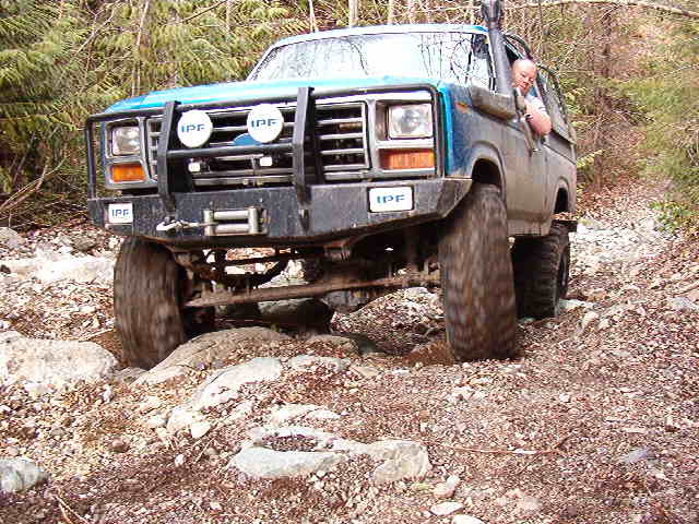 Blue 1980s Ford Bronco off-roading on a rocky trail with trees.
