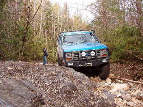 1985 Ford Bronco - A blue 1980s Ford Bronco is navigating a rocky, wooded trail with a person standing to the left of the vehicle. The Bronco has a black afte…