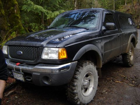 1985 Ford Bronco - A black Ford Ranger pickup truck with a camper shell is parked on a wet, muddy forest road. The truck has off-road tires and a license plat…