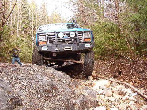 1985 Ford Bronco - A lifted, teal 1980s Ford Bronco is navigating a rocky, wooded trail. A person stands to the left of the vehicle, observing its progress.