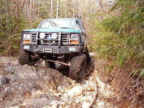 1985 Ford Bronco - A teal 1985 Ford Bronco is navigating a rocky, wooded trail. The off-road vehicle features a heavy-duty front bumper with auxiliary lights…