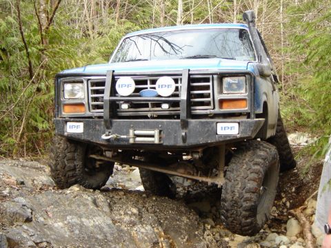 1985 Ford Bronco - A blue Ford Bronco is navigating a rocky, wooded trail with large off-road tires and a winch on the front bumper. The vehicle has IPF light…
