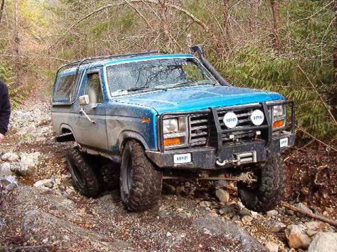 1985 Ford Bronco - A blue Ford Bronco is shown off-roading on a rocky, wooded trail. The vehicle has a custom front bumper with auxiliary lights and a winch,…