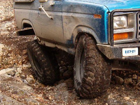 1985 Ford Bronco - The front right side of a blue, mud-covered 1980s Ford Bronco is shown at a low angle, navigating a rocky, off-road terrain. The vehicle fe…