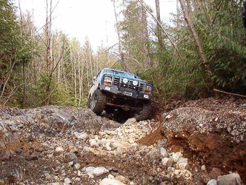 1985 Ford Bronco - A teal Ford Bronco is navigating a rocky, off-road trail surrounded by trees. The vehicle is angled upwards, with its front tires on a rock…
