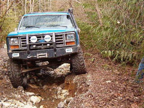 1985 Ford Bronco - A blue 1980s Ford Bronco is navigating a rocky, off-road trail with its front tires angled into a ditch. The vehicle features a brush guard…