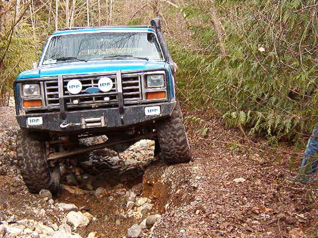 Blue 1980s Ford Bronco off-roading on a rocky trail.