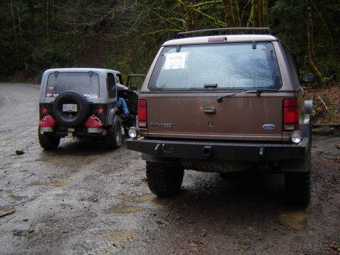 1985 Ford Bronco - The rear view of a brown Ford Explorer is shown parked on a muddy dirt road in a forest. To the left, the rear of a gray Jeep Wrangler is v…