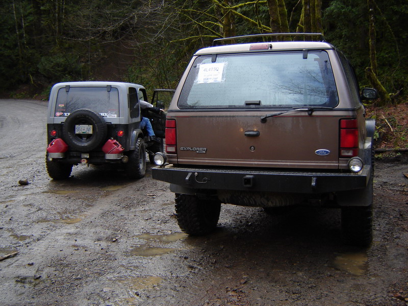 Rear view of a brown Ford Explorer and a gray Jeep Wrangler on a muddy forest road.