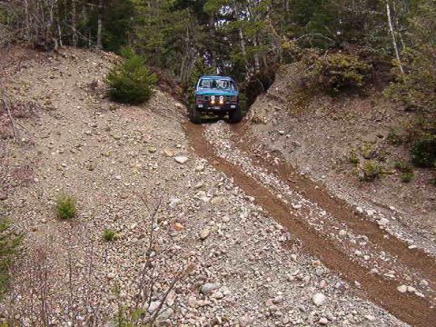 1985 Ford Bronco - A blue 1985 Ford Bronco is driving up a steep, rocky, and dirt path in a wooded area. The vehicle is angled towards the viewer, showing its…
