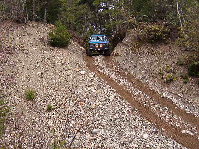 1985 Ford Bronco driving up a steep, rocky, dirt trail in a forest.