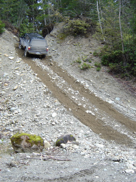 1985 Ford Bronco - A dark-colored pickup truck with a camper shell is driving down a steep, rocky, and muddy incline in a wooded area. The truck is angled awa…