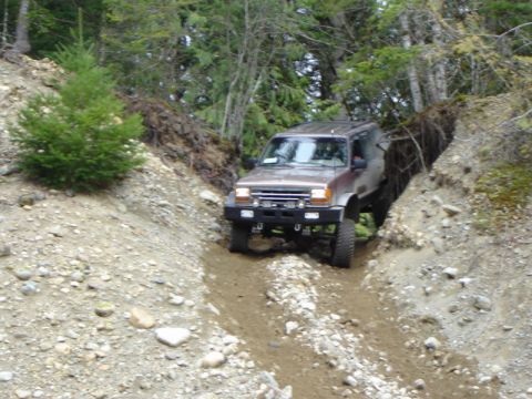 1985 Ford Bronco - A brown 1980s Ford Bronco is navigating a steep, narrow dirt trail with steep, wooded banks on either side. The vehicle is angled downwards…