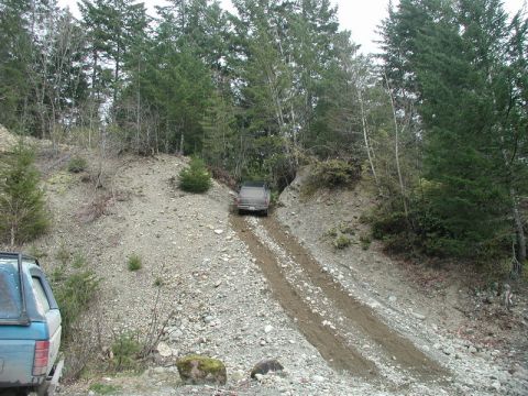 1985 Ford Bronco - A dark-colored pickup truck is driving up a steep, rocky, and muddy incline in a wooded area. Another vehicle, partially visible on the lef…