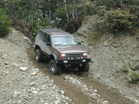 1985 Ford Bronco - A lifted, brown Ford Explorer is navigating a steep, rocky, and muddy trail in a wooded area. The SUV features an aftermarket front bumper…