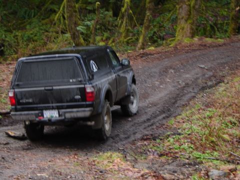 1985 Ford Bronco - A black pickup truck with a camper shell is driving on a muddy dirt road through a forest.