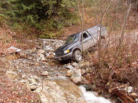 1985 Ford Bronco - A black SUV is driving through a rocky stream bed with trees and brush on either side. The vehicle appears to be tilted to the right as it…
