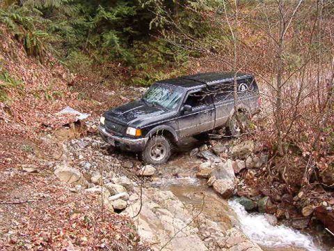 1985 Ford Bronco - A black Ford Ranger pickup truck with a camper shell navigates a rocky, wooded stream bed. The truck is angled towards the right, with its…