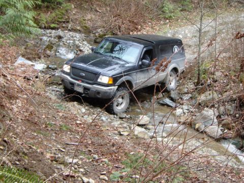 1985 Ford Bronco - A black Ford Ranger pickup truck with a camper shell is driving through a shallow rocky stream in a wooded area.