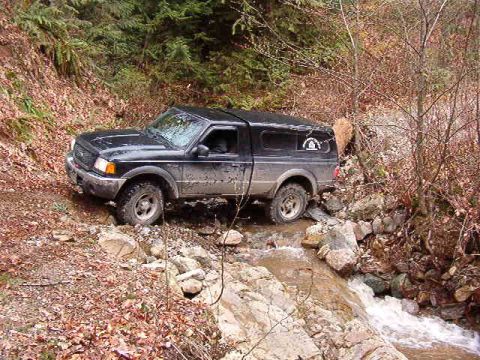 1985 Ford Bronco - A black Ford pickup truck with a camper shell drives through a rocky stream on a dirt road, surrounded by trees and fallen leaves.
