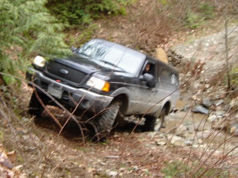 1985 Ford Bronco - A black Ford pickup truck with a camper shell is driving through a shallow stream in a wooded area. The truck is angled to the left and app…