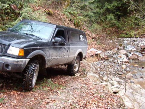 1985 Ford Bronco - A black Ford pickup truck with a camper shell is parked on a rocky, wooded trail next to a stream. The truck has off-road tires and appears…