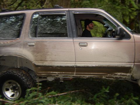 1985 Ford Bronco - Side view of a tan SUV covered in mud, driving through a forest with a person wearing a black beanie visible in the driver's seat.