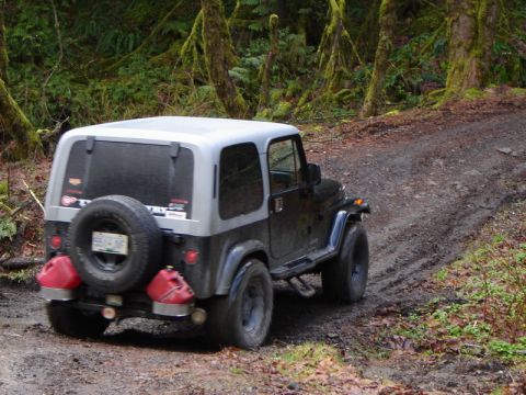 1985 Ford Bronco - A dark-colored Jeep Wrangler with a silver hardtop drives up a muddy, wooded trail. The vehicle has two red jerry cans mounted on the rear…