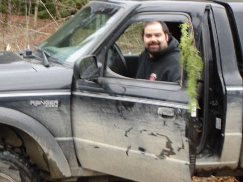 1985 Ford Bronco - A man smiles from the driver's seat of a muddy black Ford Ranger pickup truck parked in a wooded area. A branch of greenery hangs from the…