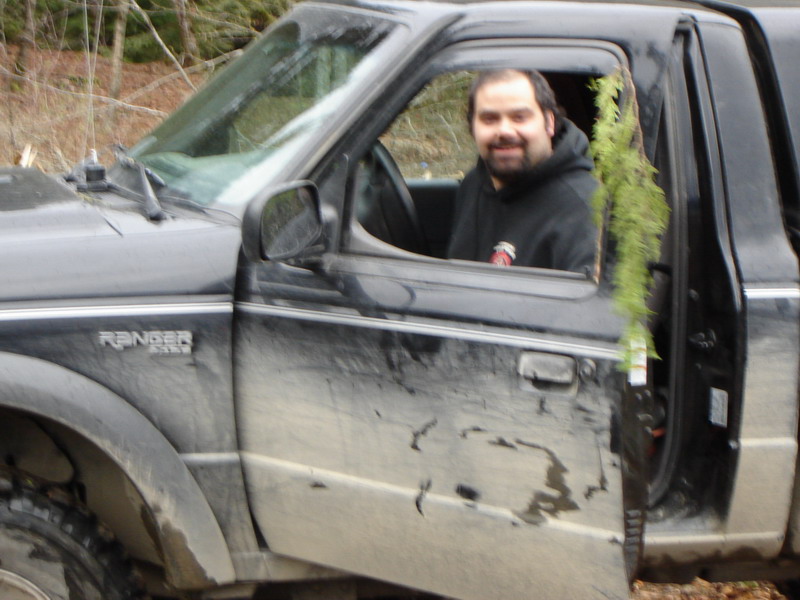 Man in a black hoodie smiles in a muddy black Ford Ranger pickup truck.