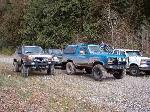 1985 Ford Bronco - Three modified off-road vehicles are parked on a gravel lot with trees in the background. A blue Ford Bronco is in the center, with a silve…