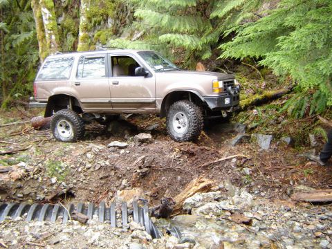 1985 Ford Bronco - A lifted, tan Ford Explorer is navigating a muddy, wooded trail with a stream and culvert in the foreground.