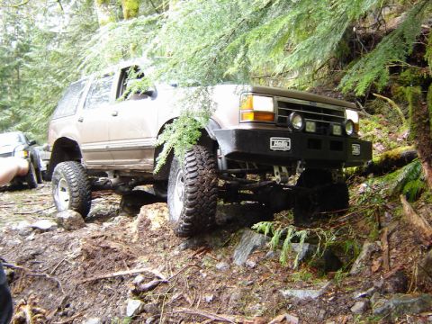 1985 Ford Bronco - A lifted, brown SUV with aggressive off-road tires navigates a rocky, wooded trail. The vehicle has a custom front bumper with Hella lights…