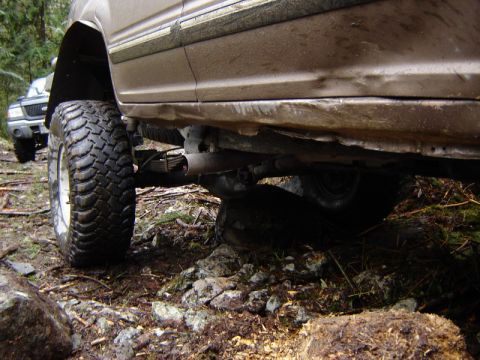 1985 Ford Bronco - The undercarriage of a lifted off-road vehicle is shown navigating a rocky, wooded terrain. The large, aggressive tires and visible suspens…