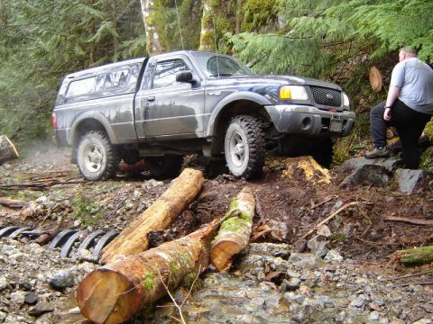 1985 Ford Bronco - A dark-colored pickup truck with a camper shell navigates an off-road obstacle course of logs and rocks in a forest setting. A person stand…