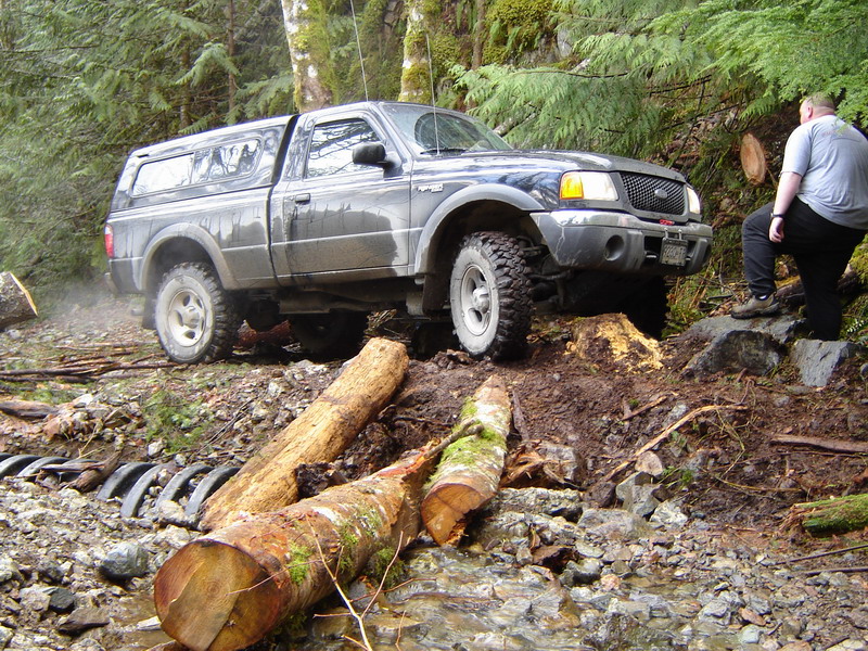 Dark pickup truck with camper shell driving over logs and rocks on an off-road trail.