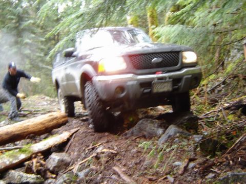 1985 Ford Bronco - A dark-colored Ford pickup truck navigates a rocky, wooded trail with a person in the background.